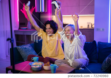 Smiling couple holding gamepads playing video game at home. Low angle shot of young people spending time together during self isolation on pandemic. Smiling couple holding gamepads playing video game at home. Low angle shot of young people spending time together during self isolation on pandemic. 72240960