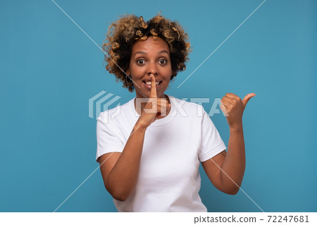 Attractive cheerful african woman pointing aside, holding finger on mouth. 72247681