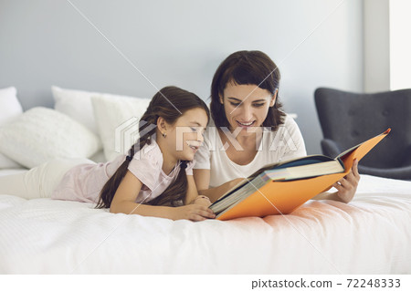 Happy mother and daughter lying together on bed and reading book 72248333
