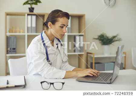 erious female practitioner typing on laptop keyword sitting at desk in office of modern clinic erious female practitioner typing on laptop keyword sitting at desk in office of modern clinic 72248359