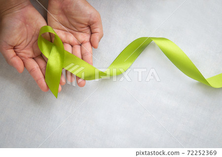 Hands holding light lime green ribbon on white fabric background with copy space. World Mental Health Day and Lymphoma Awareness symbol. Healthcare medical and insurance concept. 72252369