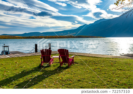 Red chair look over Middle Waterton Lake boat launch in autumn foliage season morning. Sunlight passing blue sky and clouds on mountains. Waterton Lakes National Park, Alberta, Canada. 72257310