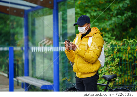 Young man with face mask on bus stop outdoors in town, using smertphone. Coronavirus concept. 72265353