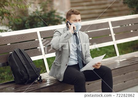 Businessman in a mask sitting on a bench with documents Businessman in a mask sitting on a bench with documents 72282862