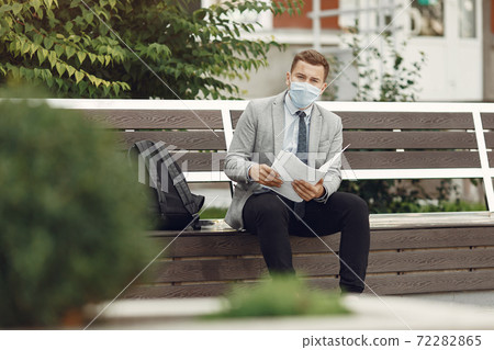 Businessman in a mask sitting on a bench with documents Businessman in a mask sitting on a bench with documents 72282865