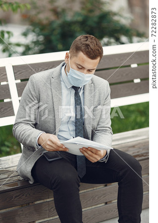 Businessman in a mask sitting on a bench with documents Businessman in a mask sitting on a bench with documents 72282873