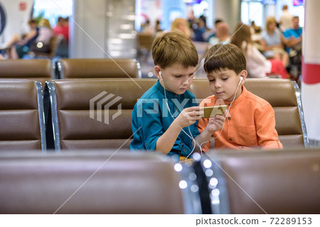Two little boy sitting in an airport departure hall contentedly playing on his tablet or mobile phone as they wait for his flight with his luggage. Children are laughing and use earphones. Travel with 72289153