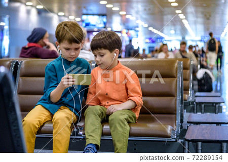 Two little boy sitting in an airport departure hall contentedly playing on his tablet or mobile phone as they wait for his flight with his luggage. Children are laughing and use earphones. Travel with Two little boy sitting in an airport departure hall contentedly playing on his tablet or mobile phone as they wait for his flight with his luggage. Children are laughing and use earphones. Travel with 72289154