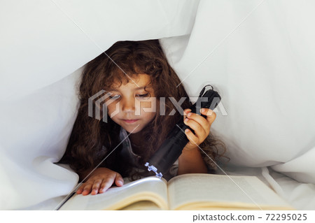 Little girl reads a book with a flashlight under a blanket in bed 72295025