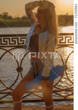 Side view of overjoyed lady with pennyboard in her hands in front of the setting sun over the water surface of the river 72299861