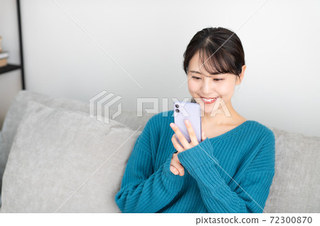 一個年輕的女人坐在客廳裡的沙發上，使用智能手機 72300870