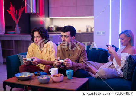 Guys playing video game while girl using smartphone for social media. Friends spending time together at home. Guys playing video game while girl using smartphone for social media. Friends spending time together at home. 72304997