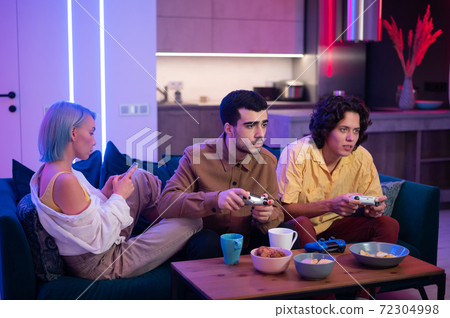 Guys playing video game while girl using smartphone for social media. Friends spending time together at home. Guys playing video game while girl using smartphone for social media. Friends spending time together at home. 72304998