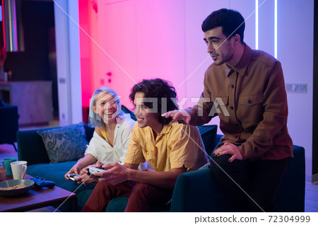 Happy young people playing video games on console while sitting on couch in front of tv. Man advice his friend in a game. Room with warm and neon lights. Happy young people playing video games on console while sitting on couch in front of tv. Man advice his friend in a game. Room with warm and neon lights. 72304999