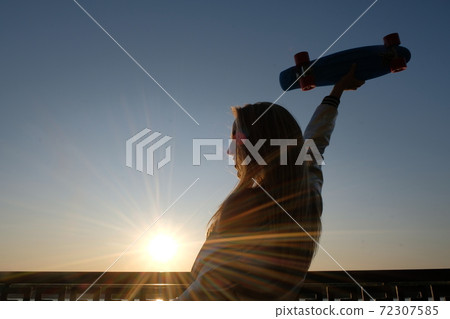 Silhouette of the extatic woman with skateboard in her raised in sunset sky hand, side view, vintage color. 72307585