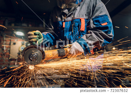 Locksmith in special clothes and goggles works in production. Metal processing with angle grinder. Sparks in metalworking 72307632