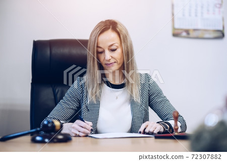 A female notary public sits at a wooden desk in her notary office signs documents. A female notary public sits at a wooden desk in her notary office signs documents. 72307882