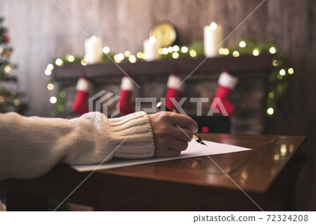 Hand of young woman with fountain pen and sheet of paper on background of christmas tree and fireplace with decoration of light bulbs. Concept of writing letter to Santa Claus 72324208