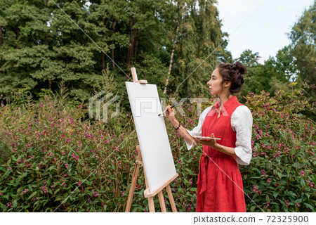 Getting started, woman in red dress, begins to paint picture, in summer on nature, background trees bushes flowers. Easel picture paint paintbrush. Free space for text. Inspiration and creativity. 72325900