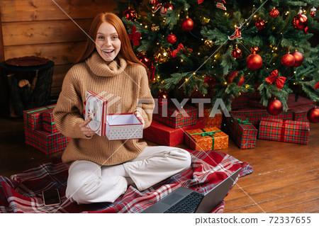 Cheerful redhead young woman opening Christmas gift box with present on background of xmas tree Cheerful redhead young woman opening Christmas gift box with present on background of xmas tree 72337655