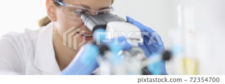 Woman in rubber gloves and protective chemical glasses looks through microscope in laboratory portrait 72354700