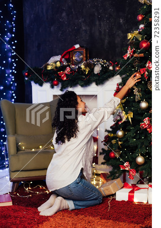 Beautiful woman decorates with toys at Christmas tree with lights garlands New Year Beautiful woman decorates with toys at Christmas tree with lights garlands New Year 72358291