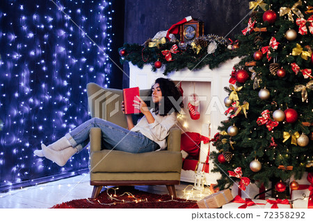 woman reads a book in a chair near the Christmas tree with lights garland new year 72358292