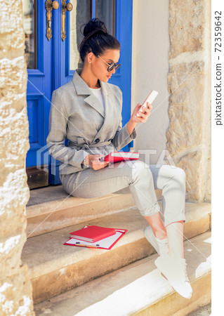 Woman in sunglasses is sitting and using smartphone near the door in the European city. 72359642