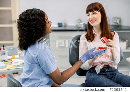 Dentist and patient in modern clinic. Smiling happy african female dentist and Caucasian girl patient holding together teeth model, speaking and looking each other. Oral care, orthodontics Dentist and patient in modern clinic. Smiling happy african female dentist and Caucasian girl patient holding together teeth model, speaking and looking each other. Oral care, orthodontics 72363241