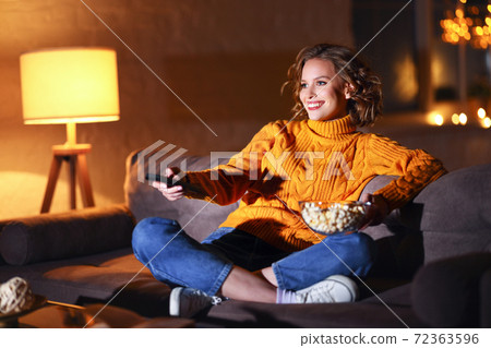 young cheerful woman eating popcorn and watches movie on TV while switching channels with remote control at home in evening alone. young cheerful woman eating popcorn and watches movie on TV while switching channels with remote control at home in evening alone. 72363596