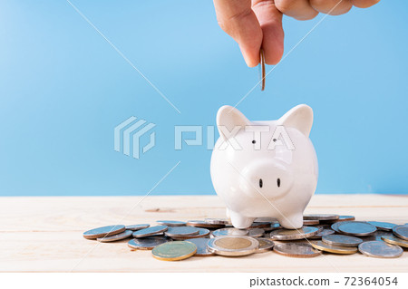 Hand putting money coin into piggy bank with a pile coins isolated blue background. Saving money and investment concept. 72364054