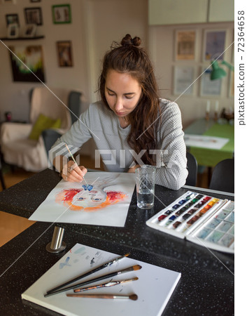 Woman at home at the table, draws paints, portrait of a girl. Bright orange paints, color palette. Black table, brush painting. Home studio creativity and art concept. Woman at home at the table, draws paints, portrait of a girl. Bright orange paints, color palette. Black table, brush painting. Home studio creativity and art concept. 72364658