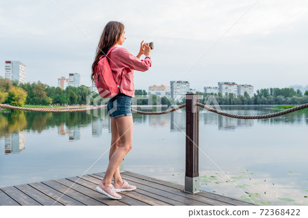 beautiful girl in a pink jacket, long hair, in summer in city, photographs lake and landscape, background of pond and river. Denim shorts on a woman. Free space for a copy of text. Green trees. 72368422