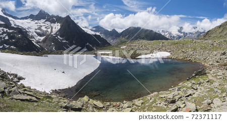 Panoramic view of glacial lake with springs from melting ice glacier with sharp snow-capped mountain peaks reflecting on water surface. Tyrol, Stubai Alps, Austria, summer sunny day 72371117