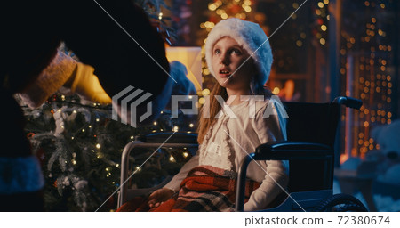 Astonished girl on wheelchair receiving present from Santa Claus at night Astonished girl on wheelchair receiving present from Santa Claus at night 72380674