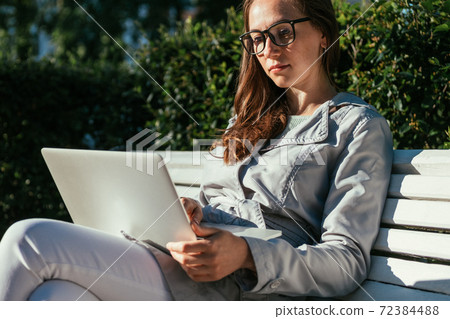 Caucasian student girl sitting on a park bench with a laptop and studying remotely. 72384488