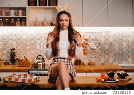 A girl with reindeer horns at Christmas sits on the kitchen table and smiles.Woman in new year's in the kitchen A girl with reindeer horns at Christmas sits on the kitchen table and smiles.Woman in new year's in the kitchen 72388158