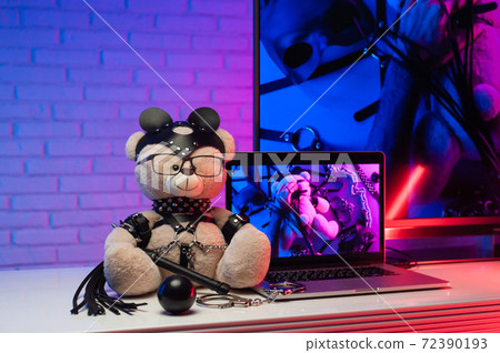 toy bear in a leather belt accessory for BDSM games next to a laptop TV in neon colors 72390193