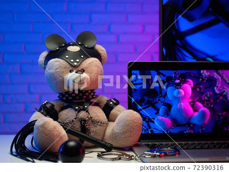 toy bear in a leather belt accessory for BDSM games next to a laptop TV in neon colors toy bear in a leather belt accessory for BDSM games next to a laptop TV in neon colors 72390316