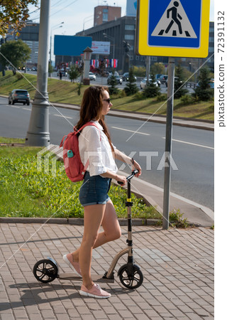 woman on scooter in summer, stands at an intersection by road, waiting to cross traffic light. Short denim shorts, white shirt and pink bag on back. woman on scooter in summer, stands at an intersection by road, waiting to cross traffic light. Short denim shorts, white shirt and pink bag on back. 72391132