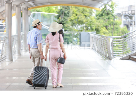senior asian traveler couple husband and wife in pink and blue shirt with white nice hat and luggage walking in downtown, they pointing something with right copy space. elderly people lifestyle 72395320