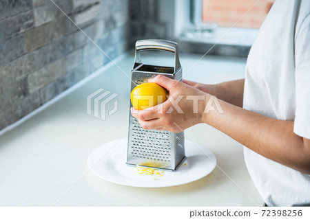 Female hands grating fresh whole lemon on metal hand grater, making zest on her kitchen table. Yellow and gray trendy colors of the year 2021. Selective focus. Copy space. Female hands grating fresh whole lemon on metal hand grater, making zest on her kitchen table. Yellow and gray trendy colors of the year 2021. Selective focus. Copy space. 72398256