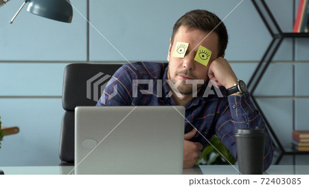 Tired comic male manager pretends working sleeping with stickers on face sits at desk with laptop. Tired comic male manager pretends working sleeping with stickers on face sits at desk with laptop. 72403085