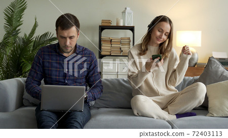 Happy couple with modern gadgets at home. A man is working on a laptop and a girl with headphones listening to music on a mobile phone is resting on the couch Happy couple with modern gadgets at home. A man is working on a laptop and a girl with headphones listening to music on a mobile phone is resting on the couch 72403113