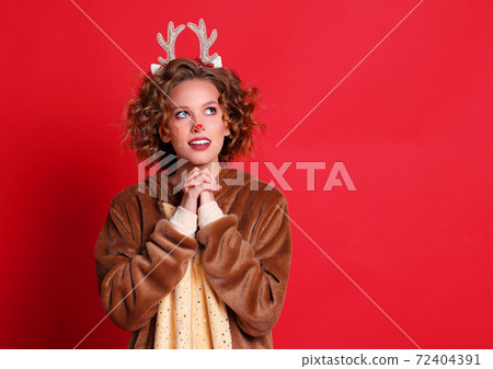 Cute young woman in a Christmas reindeer costume dreamily looks up and makes a wish on colorful red background Cute young woman in a Christmas reindeer costume dreamily looks up and makes a wish on colorful red background 72404391