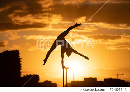 Flexible female circus Artis keep balance on one hand on the rooftop against dramatic sunset and cityscape. Handstand and equilibrium concept  72404541