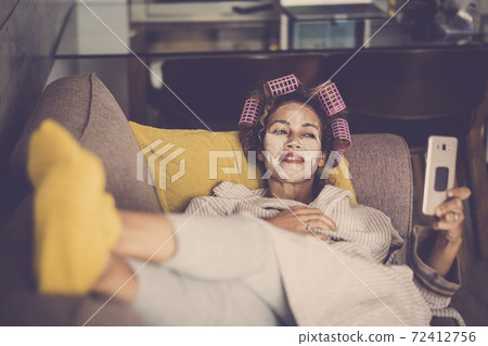 Cheerful adult woman at home enjoying time with phone video call while have a beauty mask on face and curlers on hair for beauty skin and hairstyle activity 72412756