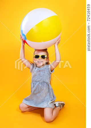 Summer girl wearing sunglasses holding ball laughing on yellow background 72416459