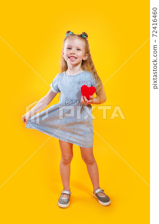 Adorable little model in trendy dress posing with red heart against yellow background 72416460
