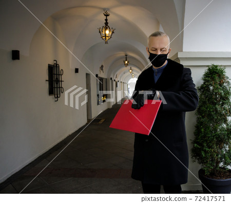 man in a black coat holds a red folder in his hands and watches the clock.  72417571